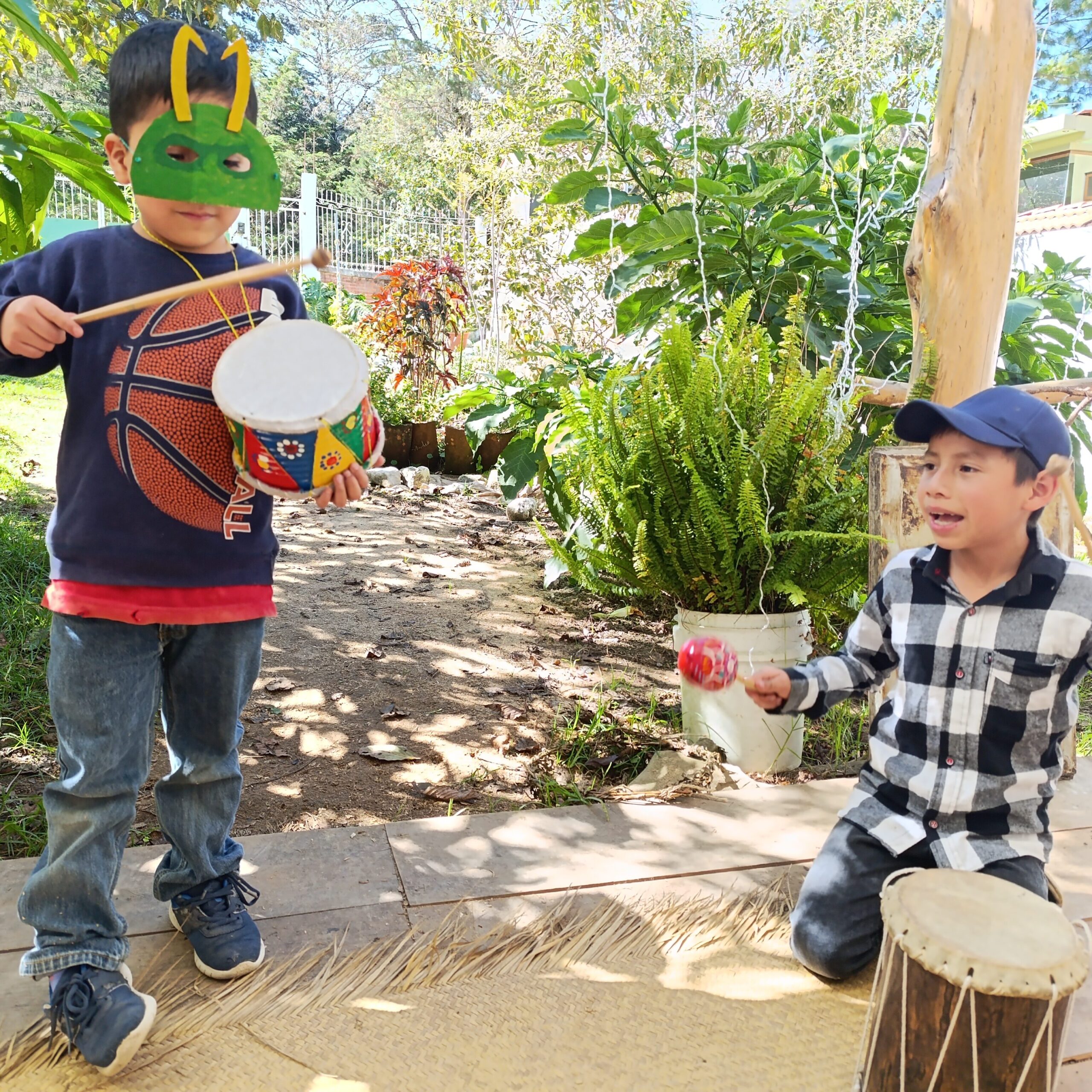 Dos niños en un jardín, uno con una máscara de insecto tocando un tambor y otro sentado con un instrumento de percusión.