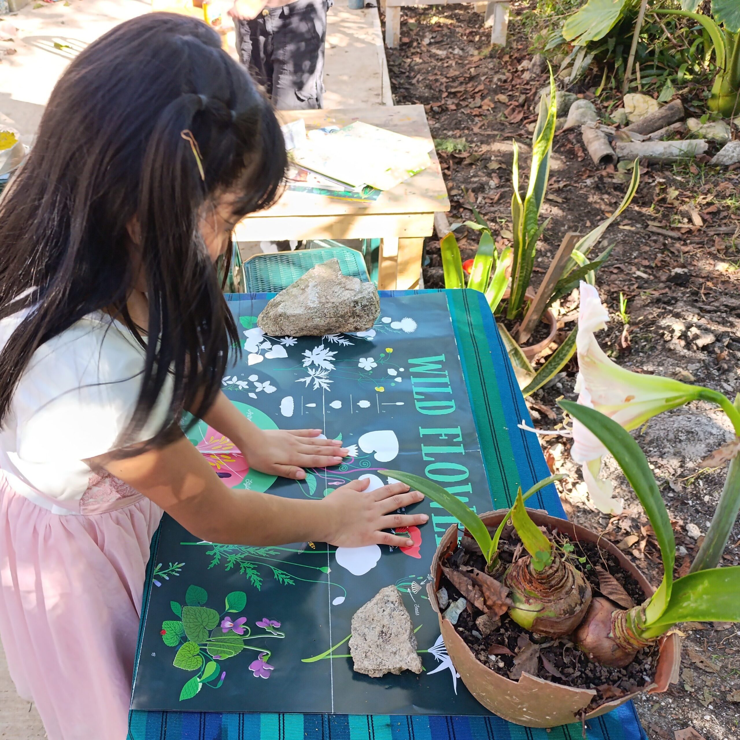 Niña trabajando en una mesa con materiales de manualidades, rodeada de plantas y piedras.